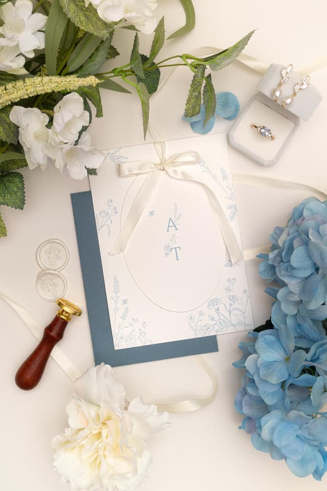Blue floral oval wedding invitation with A & T initials and fine-line flowers, tied with an ivory satin bow over a dusty blue envelope, beside ivory floral wax seals, a wax seal stamp, blue hydrangeas, white blossom and a ring box