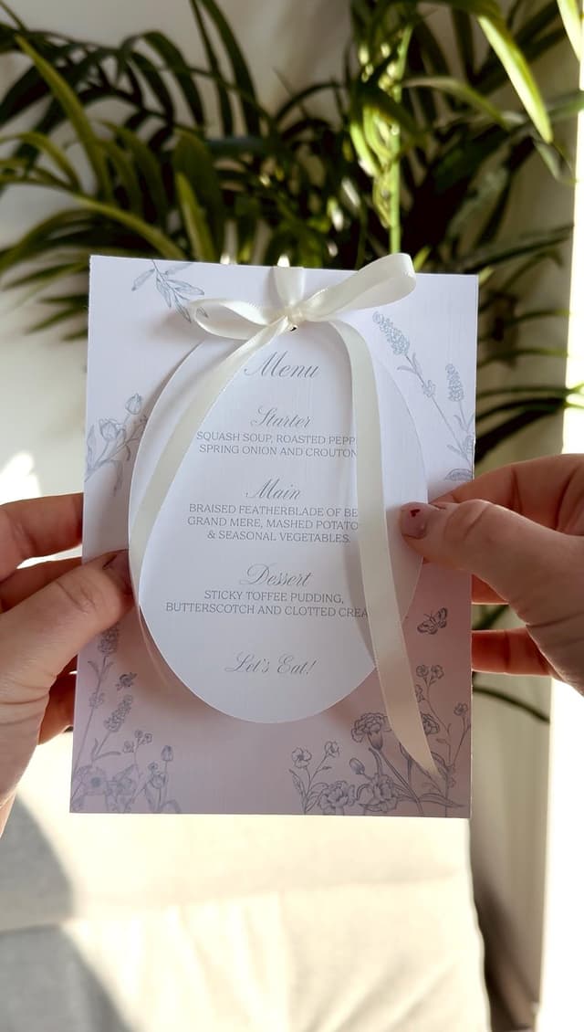 A wedding menu card with a white oval overlay, tied with a white ribbon. The menu card has a floral design in light grey. The menu includes the words "Menu", "Starter", "Main", "Dessert", and "Let's Eat!"
