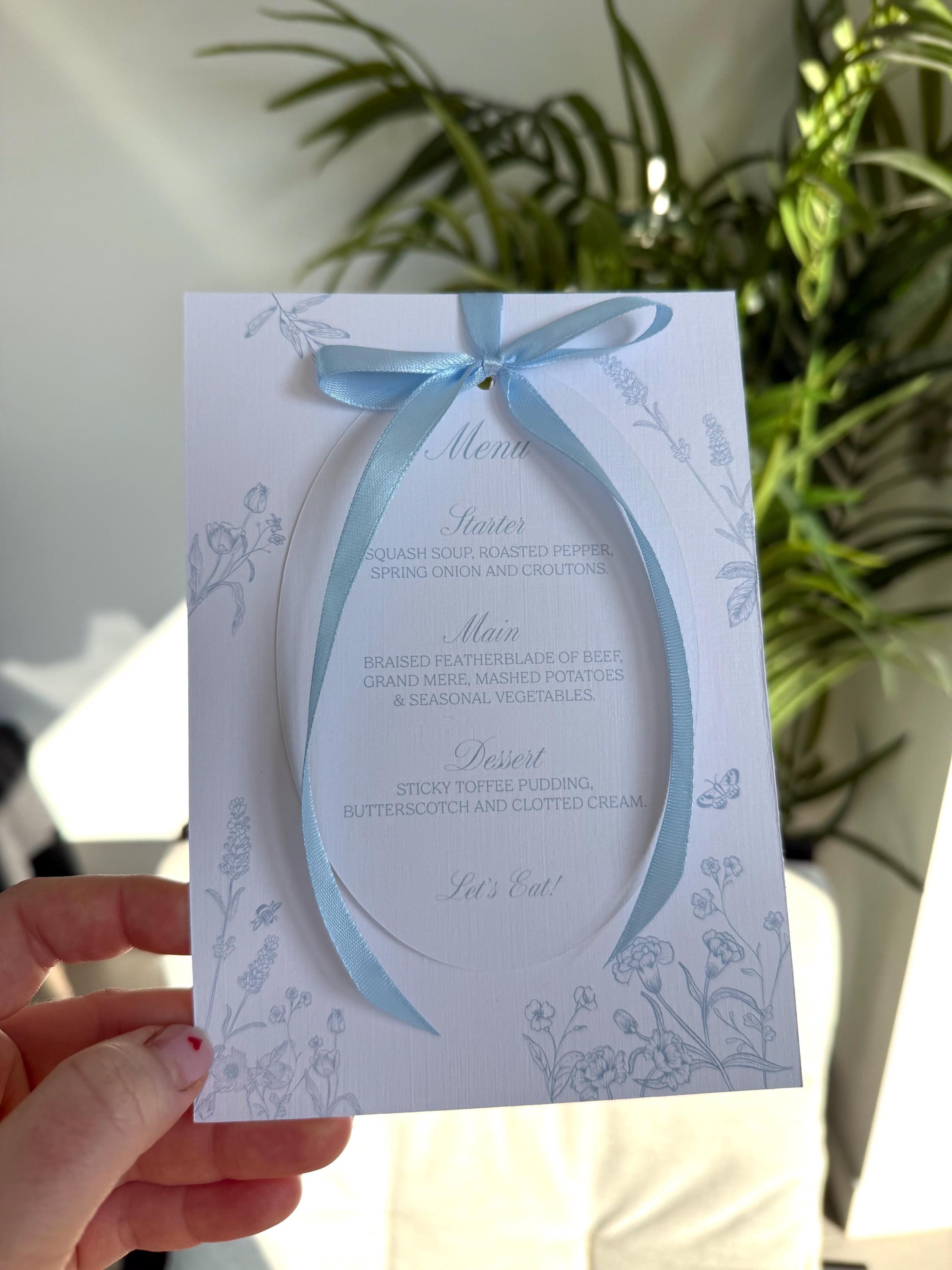 Blue floral oval wedding menu tied with a pale blue satin bow over a 5×7 card illustrated with blue wildflowers, a bee and a butterfly, ending with “Let’s Eat!”