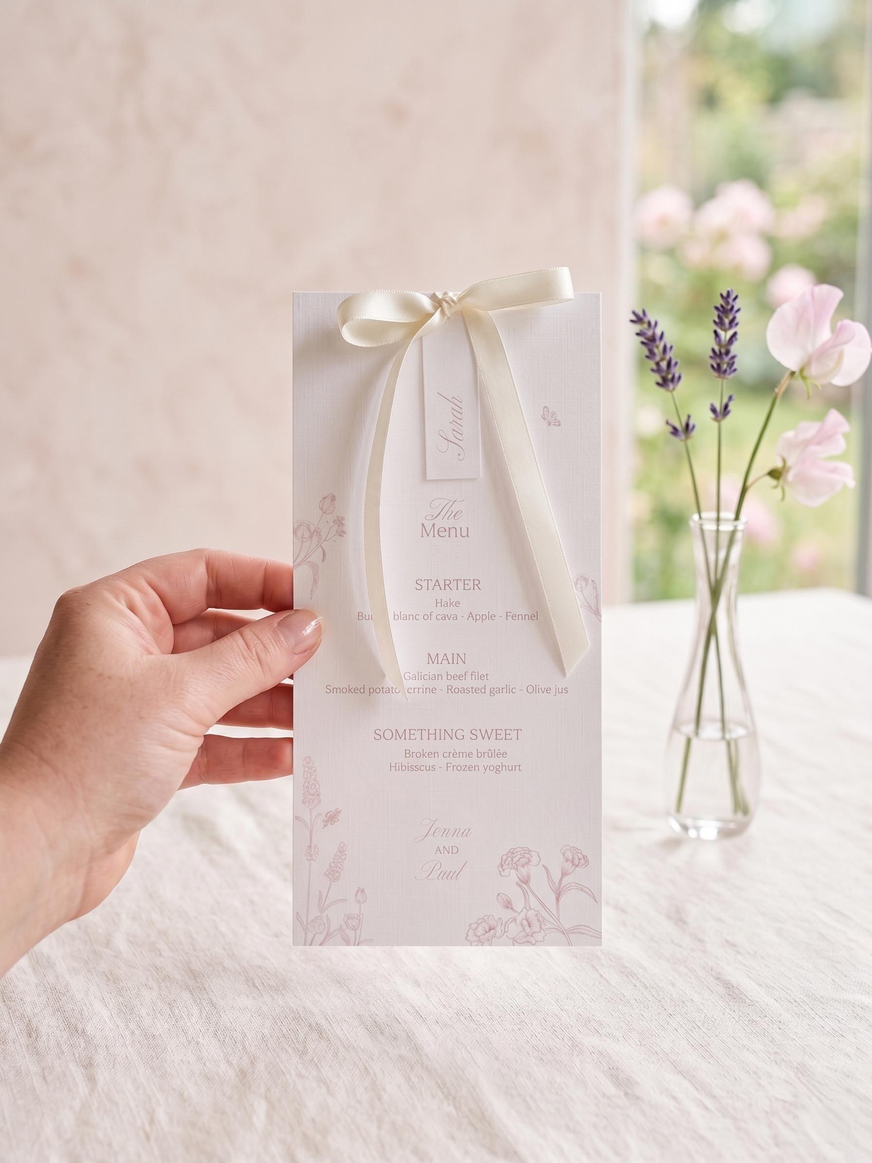 Hand holding a blush pink wildflower wedding menu for Jenna and Paul with an ivory satin bow and a name tag reading ‘Sarah’, listing a starter, main and dessert, beside a glass vase of lavender and sweet peas