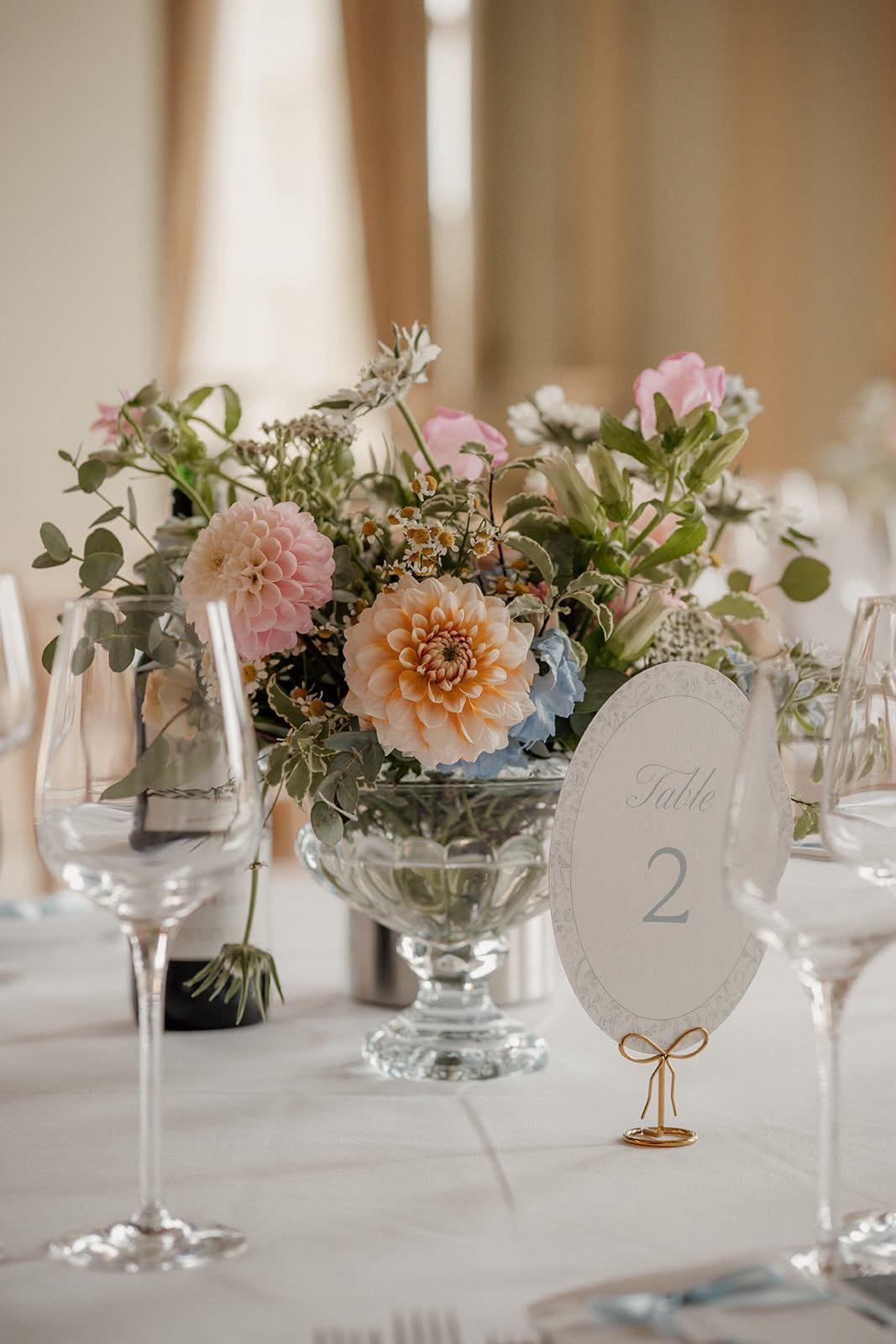 Dusty blue oval table number 2 on a gold bow-shaped stand beside a centrepiece of peach and pink dahlias, pink roses, chamomile, eucalyptus and pale blue flowers in a glass pedestal bowl