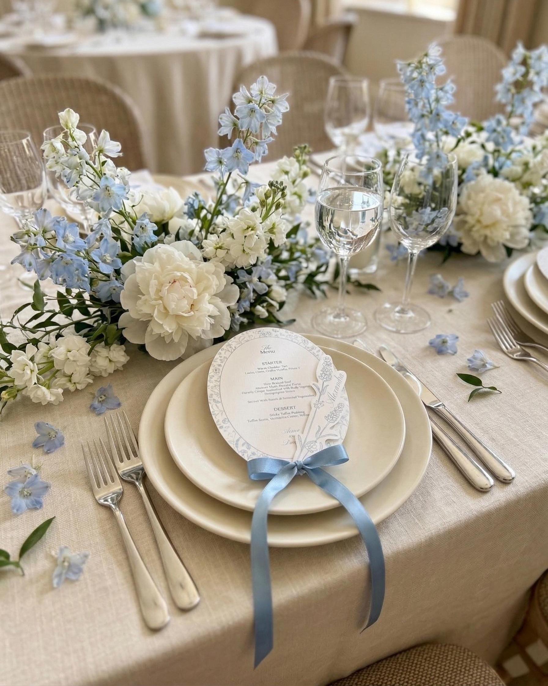 Dusty blue oval wildflower wedding menu tied with a blue satin bow on a cream place setting, with white peonies and blue delphiniums