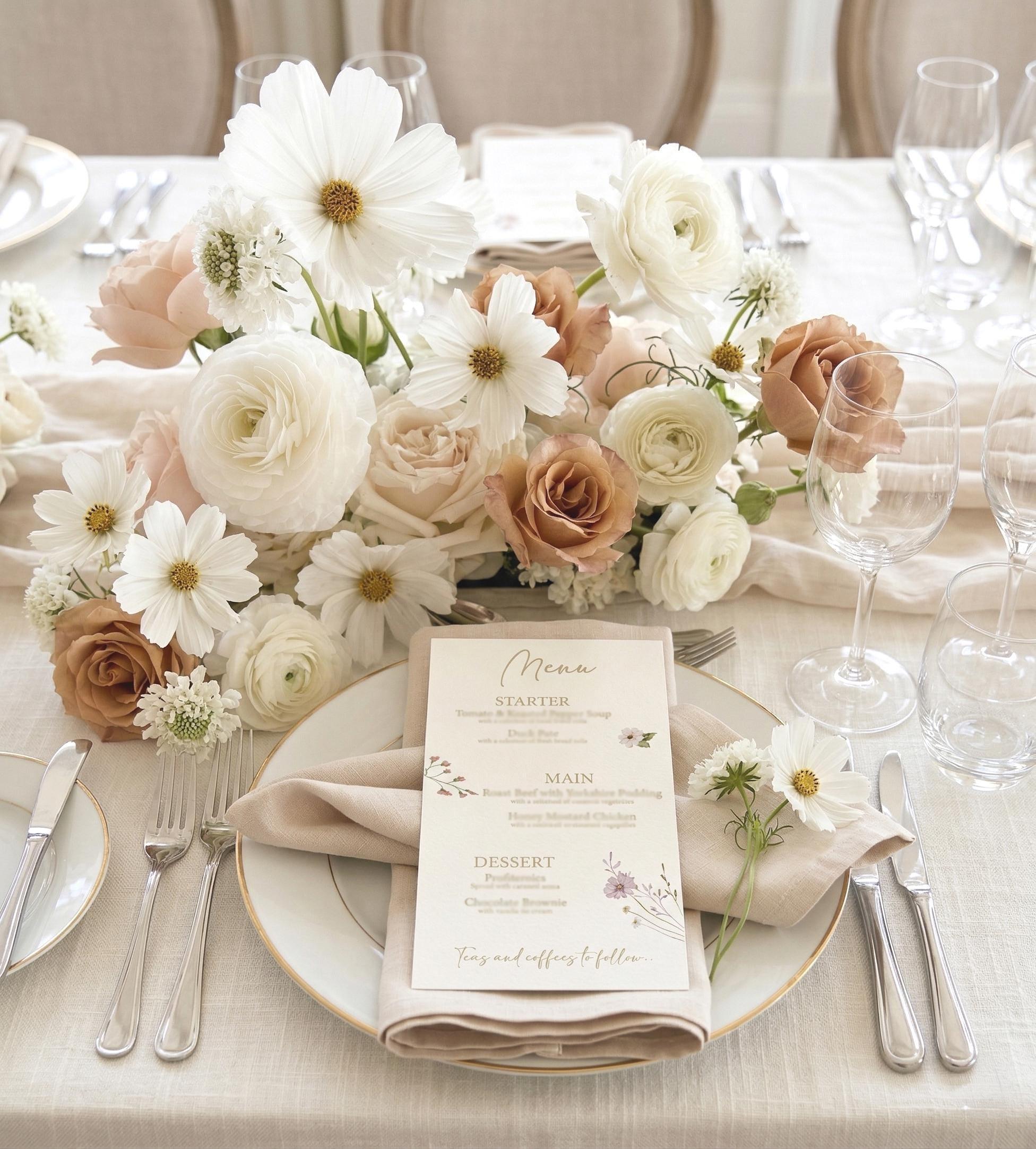 Soft floral wildflower wedding menu with two choices for each course and “Teas and coffees to follow” in gold script, on a taupe linen napkin and gold-rimmed plate beside white cosmos and caramel roses