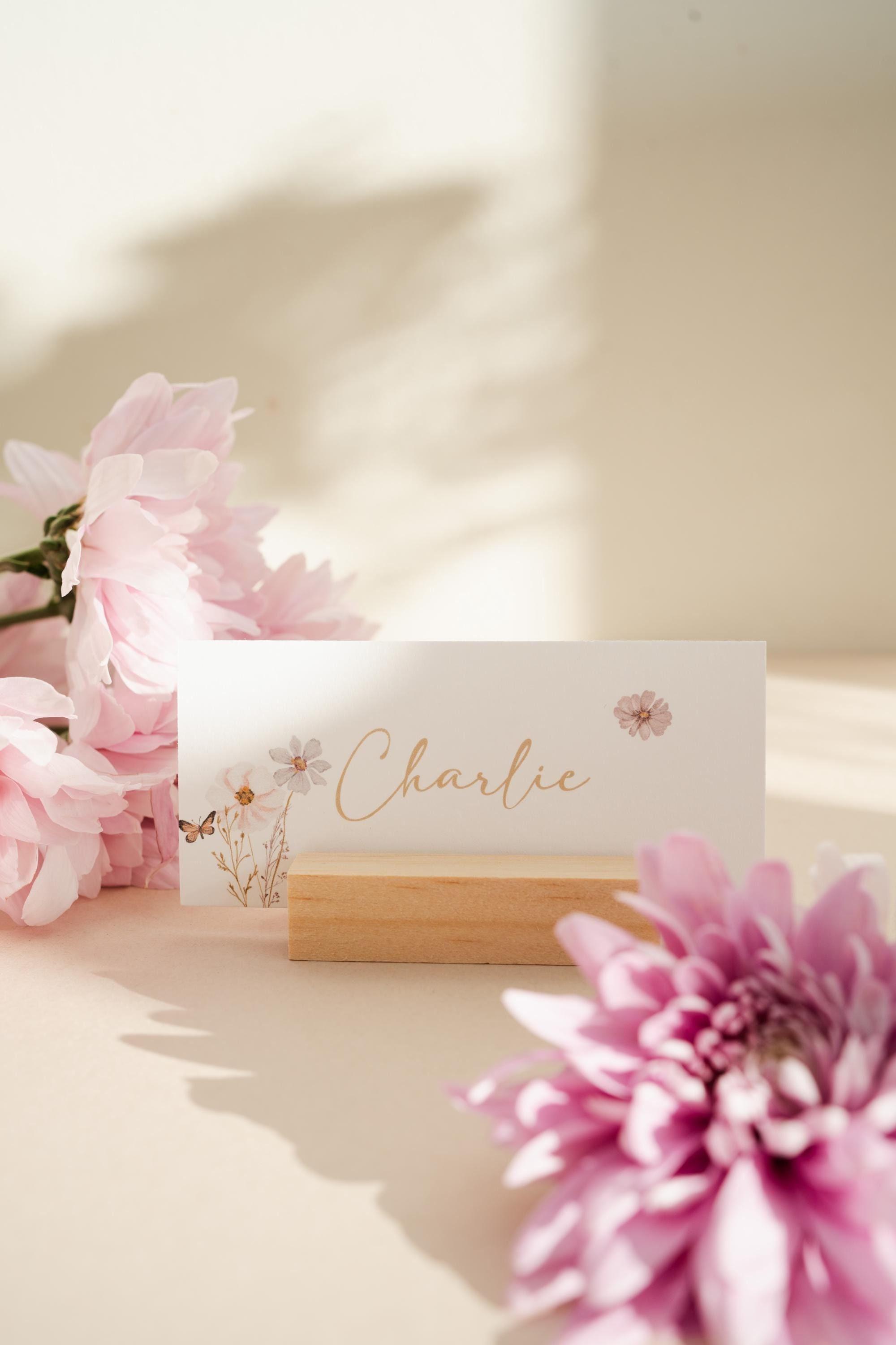 Wildflower place name card reading ‘Charlie’ in gold script with watercolour flowers and a butterfly, standing in a wooden holder beside pink chrysanthemums