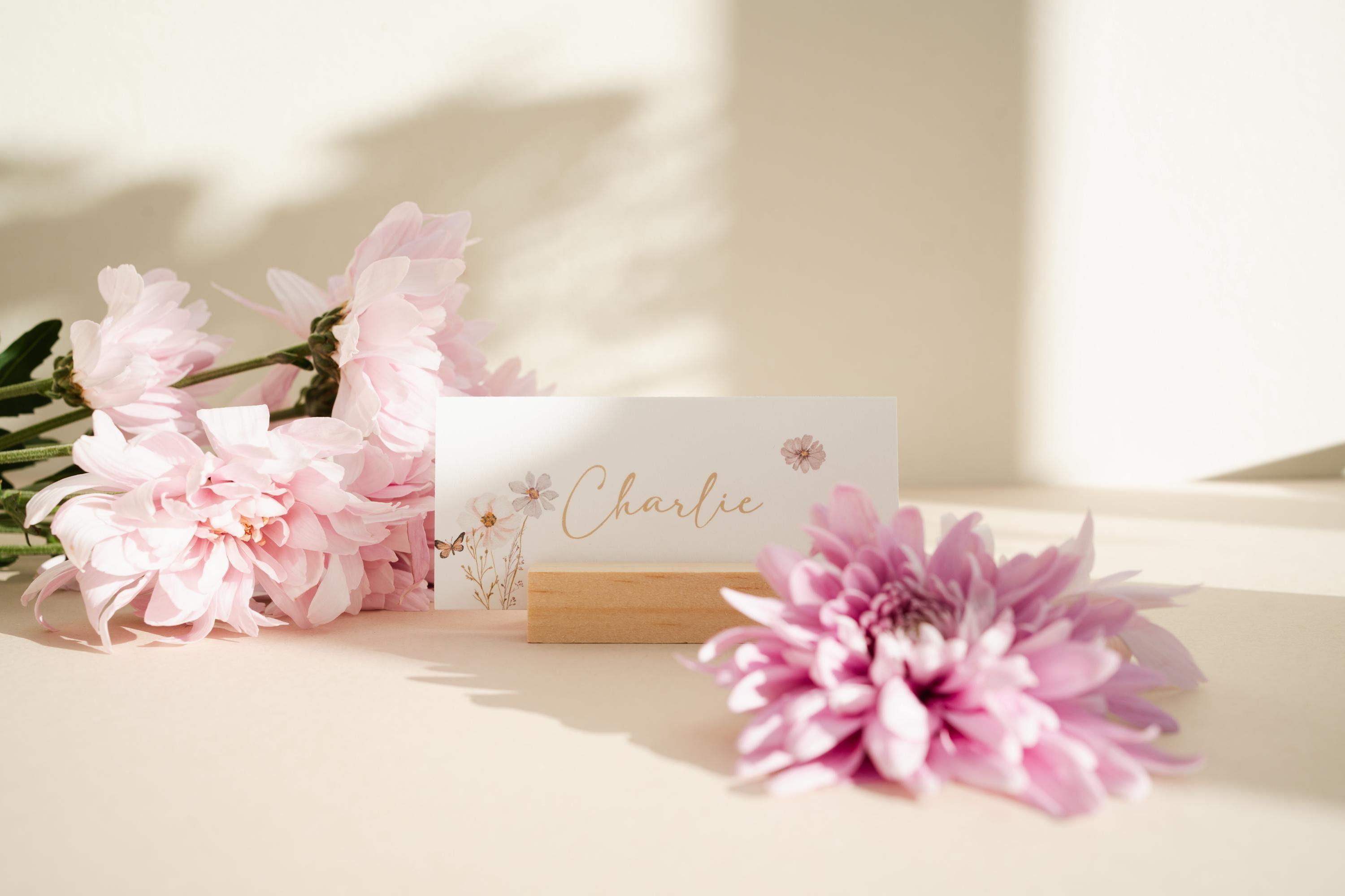 Wildflower place name card reading Charlie in gold script, with watercolour cosmos and a butterfly, standing in a wooden holder beside pink chrysanthemums