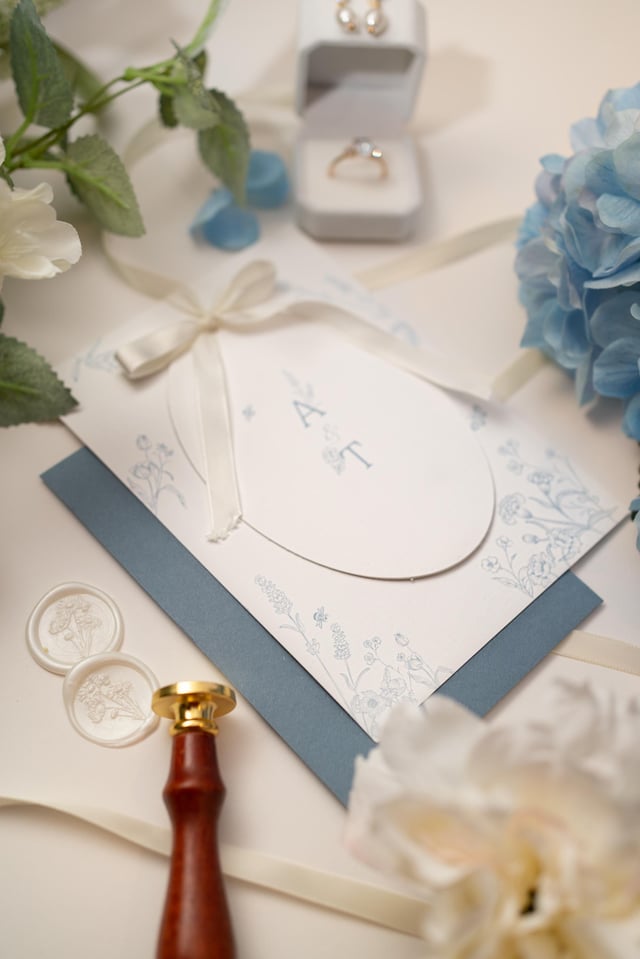 Wedding stationery set with a white invitation, blue floral design, and initials "A T". Includes a blue envelope, wax seals, and a gold ring. Decorated with blue and white flowers and ribbons.