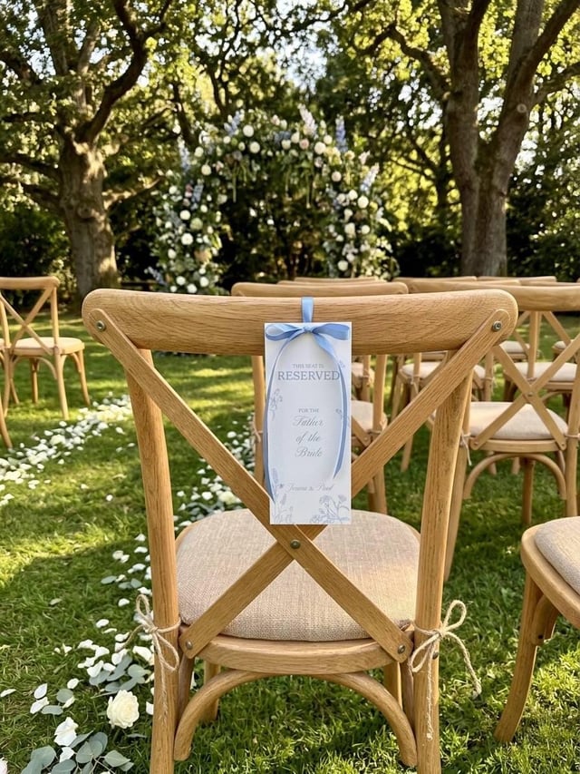 Blue wildflower reserved seat tag on an oak cross-back chair at an outdoor ceremony, with a petal-strewn aisle and a floral arch under oak trees.