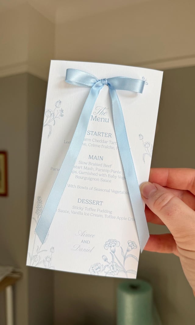 A wedding menu card tied with a light blue satin ribbon. The menu includes floral designs and the words "The Menu", "STARTER", "MAIN", and "DESSERT". The names "Aimee and Daniel" are printed at the bottom.