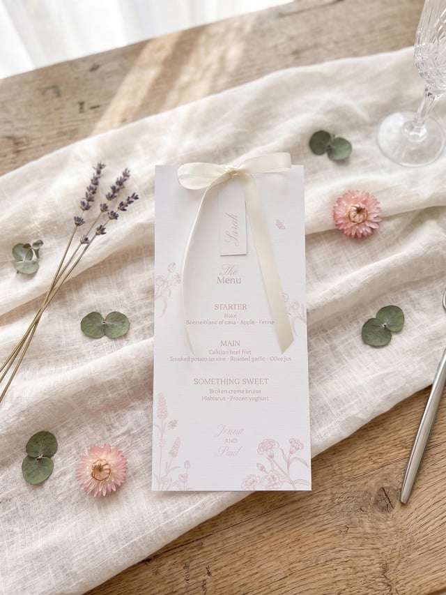 A wedding menu card with a cream ribbon and a tag that says "Sarah". The menu features floral illustrations and a list of dishes. Dried flowers and greenery are scattered around the card on a white cloth.