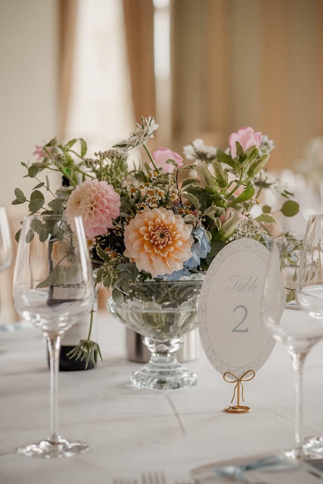 Dusty blue oval toile table number for Table 2 on a gold wire stand with a bow, beside a glass pedestal bowl of peach and pink dahlias.