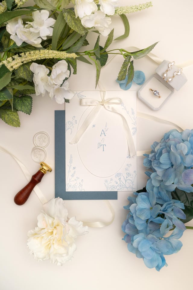 A wedding invitation featuring a blue and white floral design. The invitation is tied with a white ribbon and has the initials "A & T" in the centre. The invitation is surrounded by white flowers, blue flowers, and a wax seal.