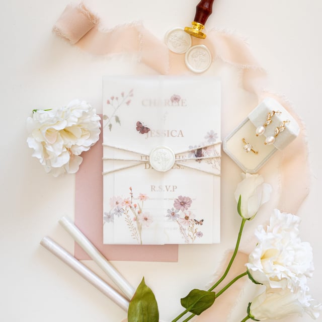 Grace wildflower invitation suite wrapped in a vellum jacket tied with twine and an ivory wax seal, on a blush envelope beside peach silk ribbon, an engagement ring and pearl earrings.