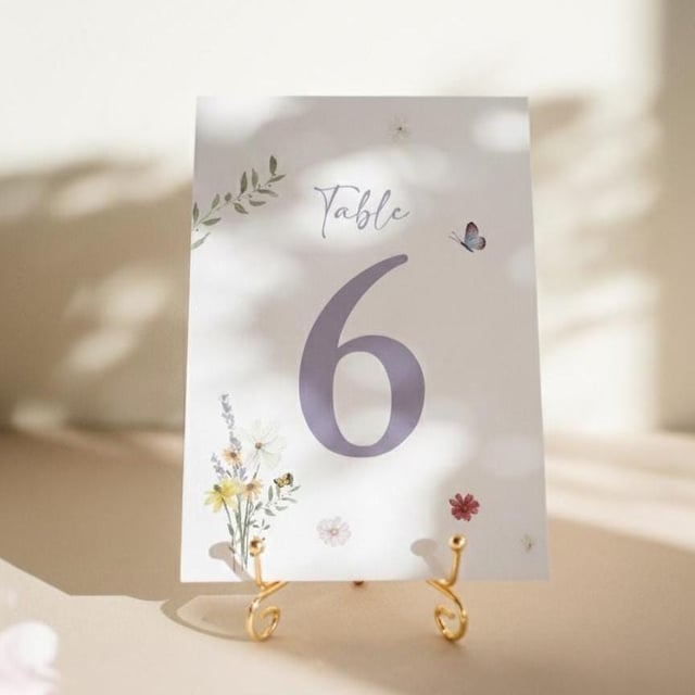 A white table number card featuring the number 6 in lavender. The card is adorned with floral and butterfly illustrations. It is supported by a gold-coloured metal stand. The word "Table" is written at the top of the card.