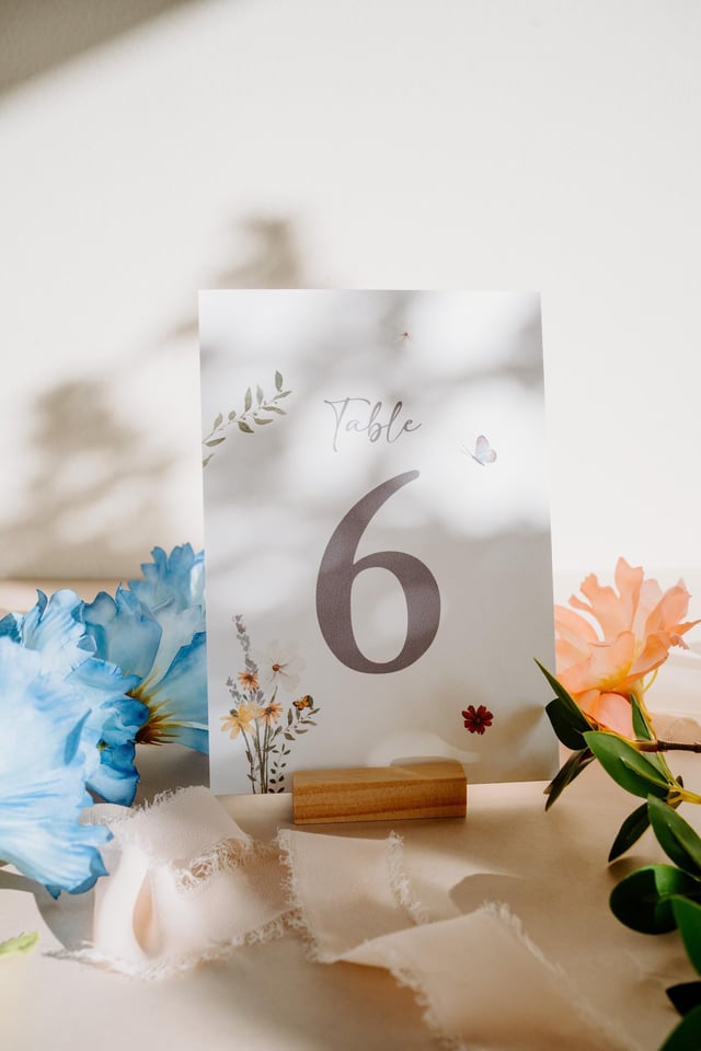 A table number card with the number 6 in a soft grey font. The word "Table" is written above the number. The card is decorated with floral illustrations and rests on a wooden block.