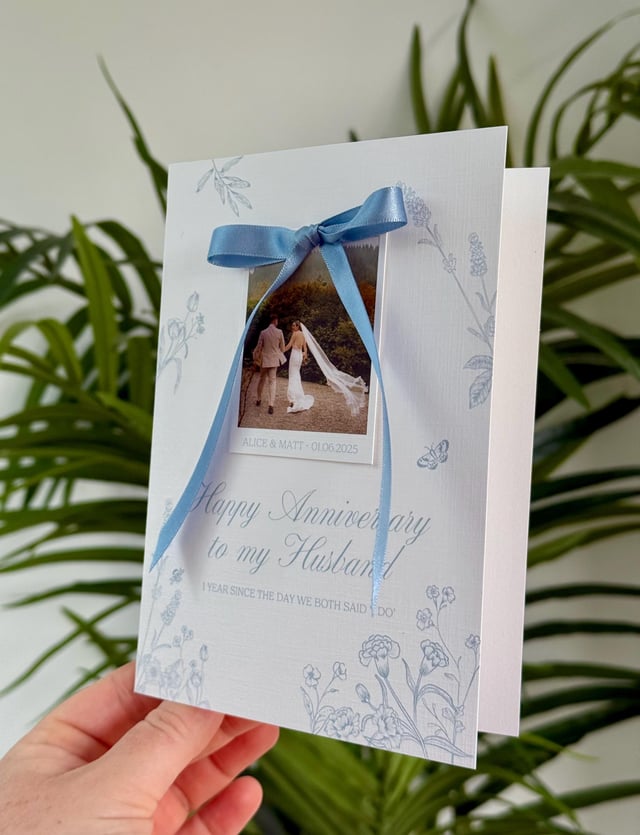A white anniversary card with a light blue ribbon and a photo of a couple. The card features floral and butterfly designs, with the text "Happy Anniversary to my Husband" and "1 year since the day we both said I do."
