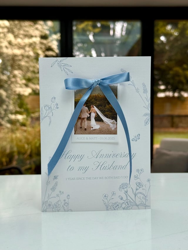 Personalised photo anniversary card with a dusty blue bow over a wedding photo of Alice & Matt, reading Happy Anniversary to my Husband, standing on a white table.