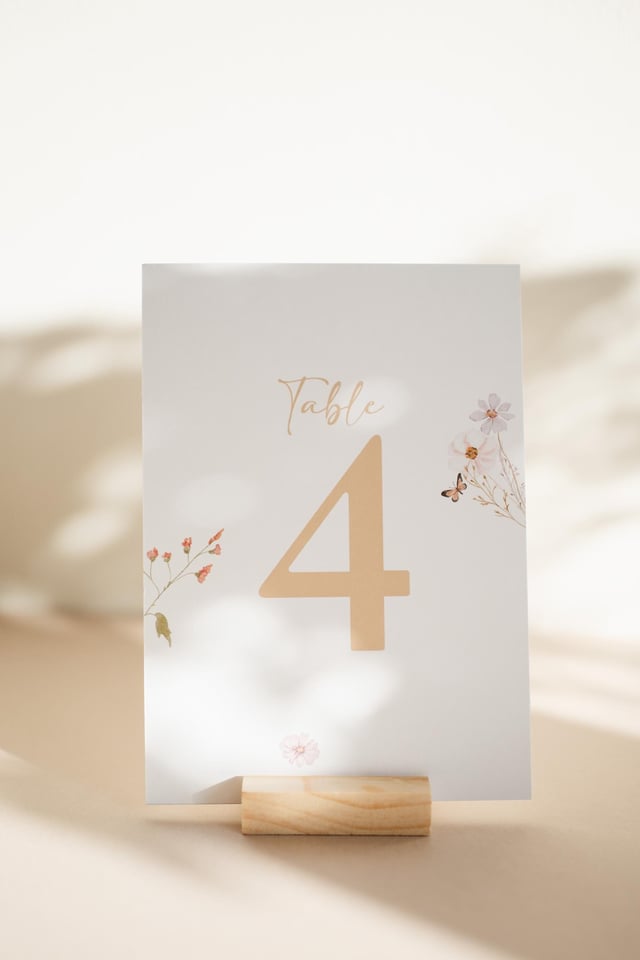 White card with the text "Table 4" in gold lettering. The card has a floral design with pink and white flowers and a butterfly. The card is on a wooden stand.