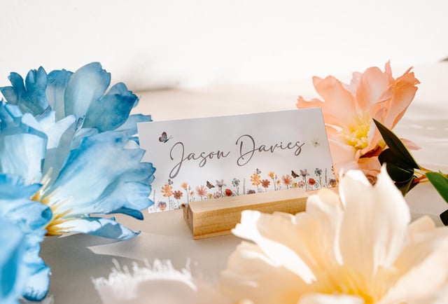 A white card with the name "Jason Davies" written in black cursive script. The card is decorated with a floral border and butterflies. The card is sitting on a wooden stand.