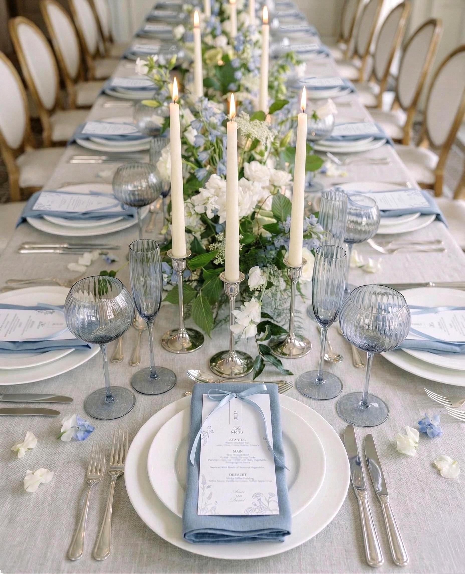 Long wedding breakfast table dressed in grey-blue linen, with dusty blue napkins, blue fluted glassware, silver candlesticks with ivory taper candles and a runner of white roses, blue delphiniums and greenery, each place set with a botanical menu tied with a pale blue ribbon